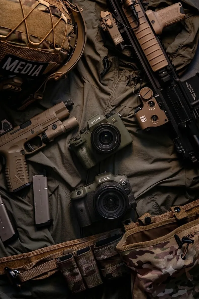 Kit de mídia em campo disposto sobre lona verde-oliva: câmeras Sony e Canon, capacete com patch MEDIA, pistola e carabina